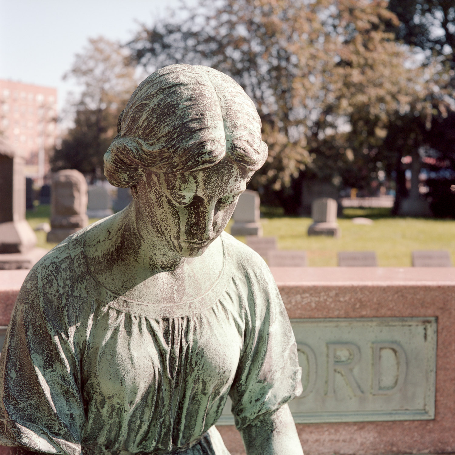 Color image of a seated bronze statuary with a heavy patina looking downwards.