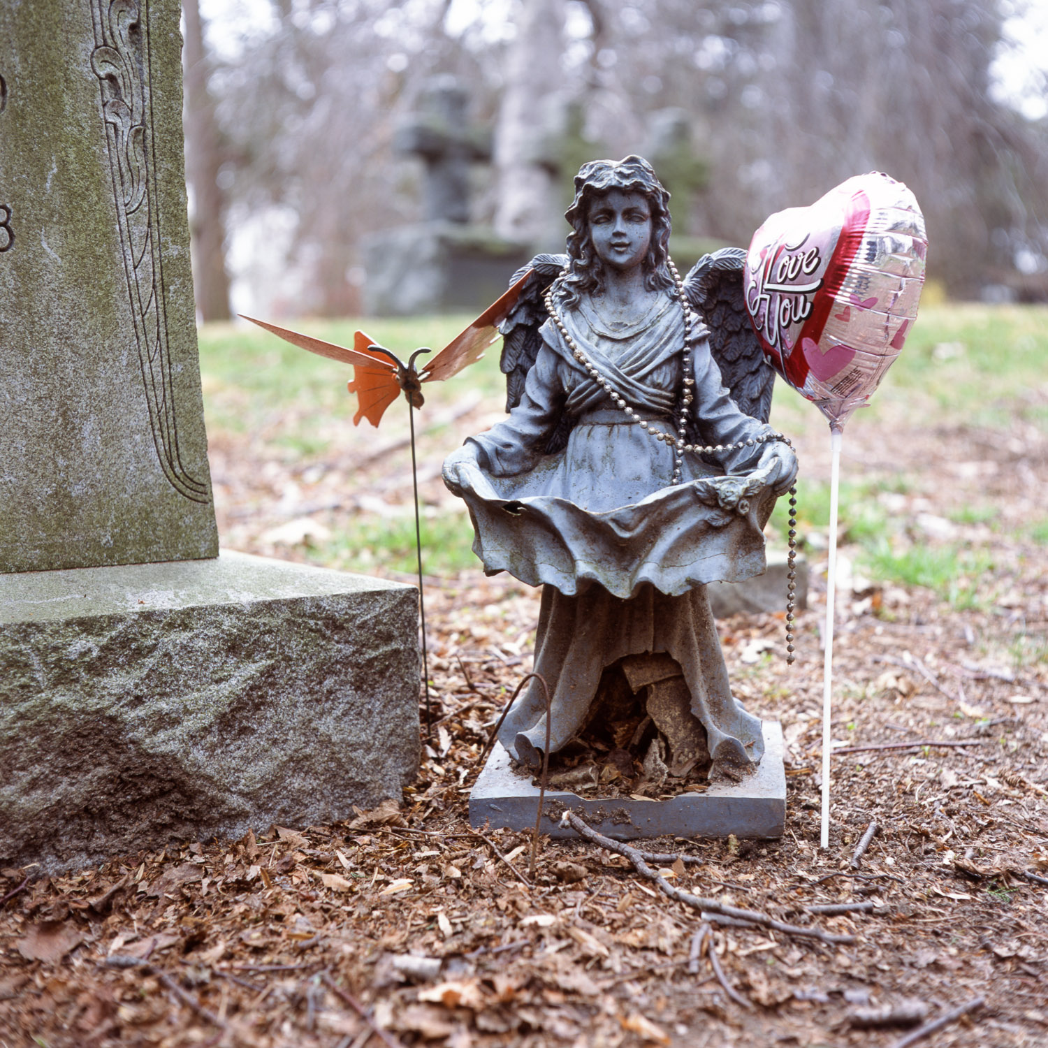 Color image of a small bronze winged statuary decorated with beads, a plastic butterfly and a 'I Love You' foil balloon next to a tombstone.