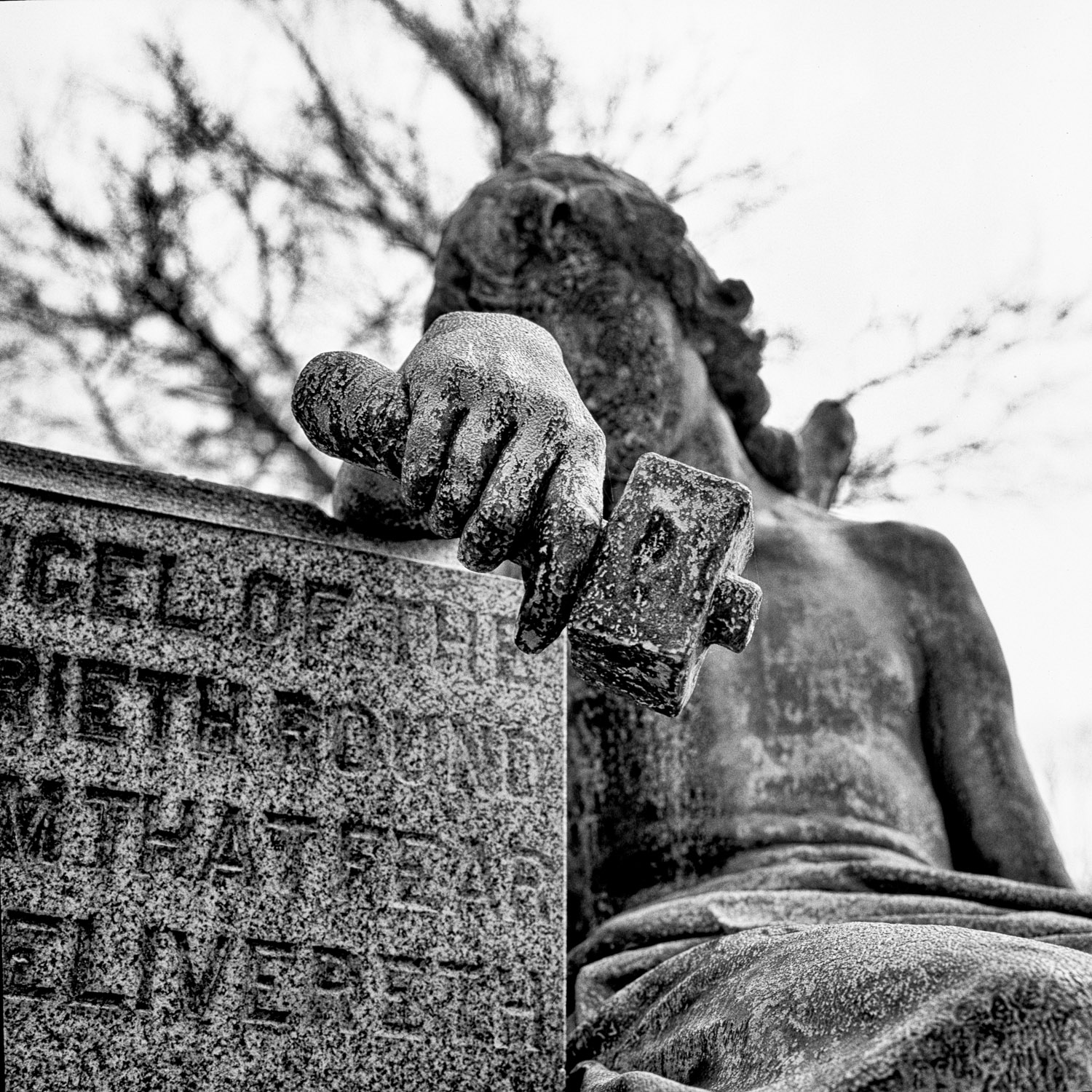 B&W image of seated stone statuary holding a hammer over a tombstone.