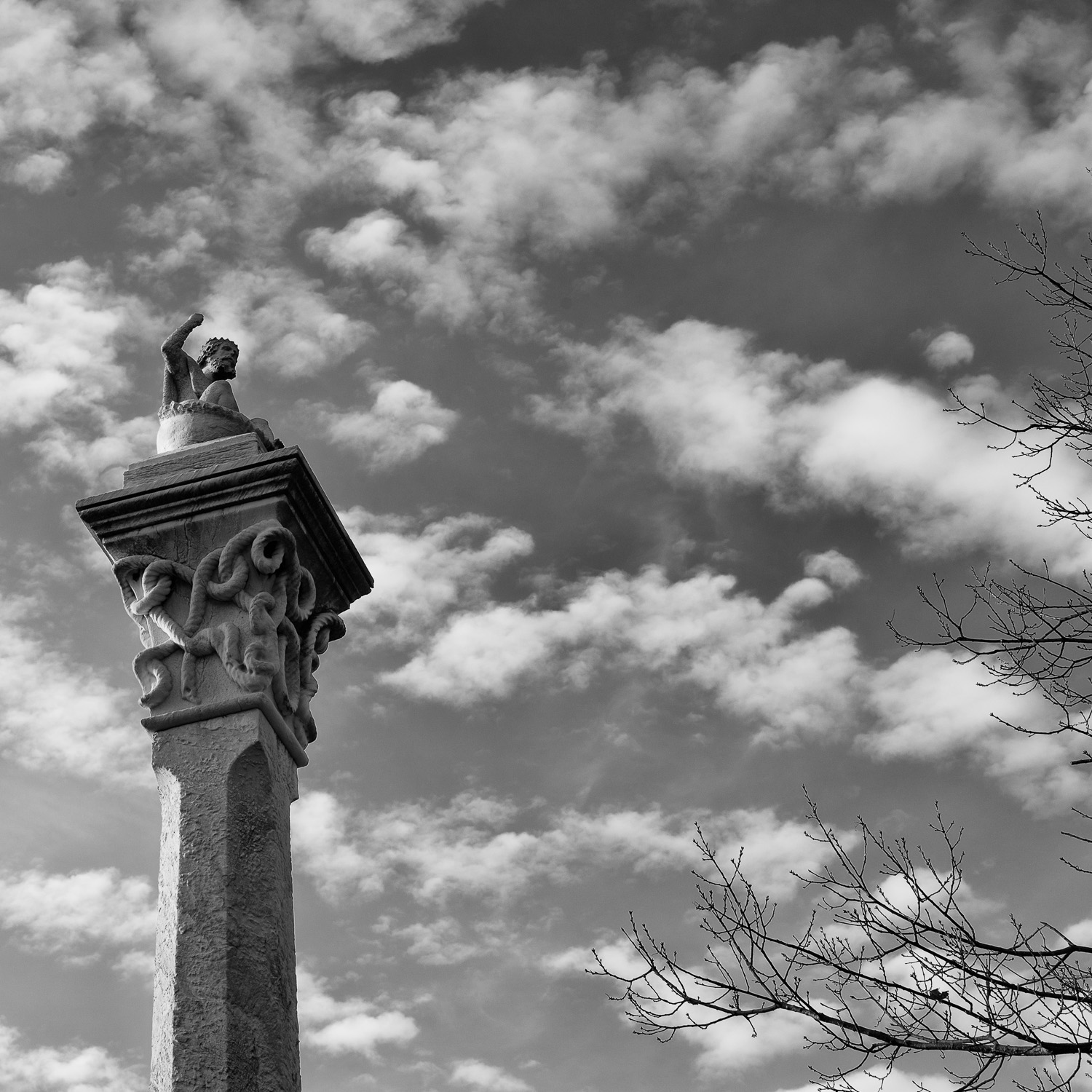 B&W image of a stone statuary with arm raised atop a tall column.