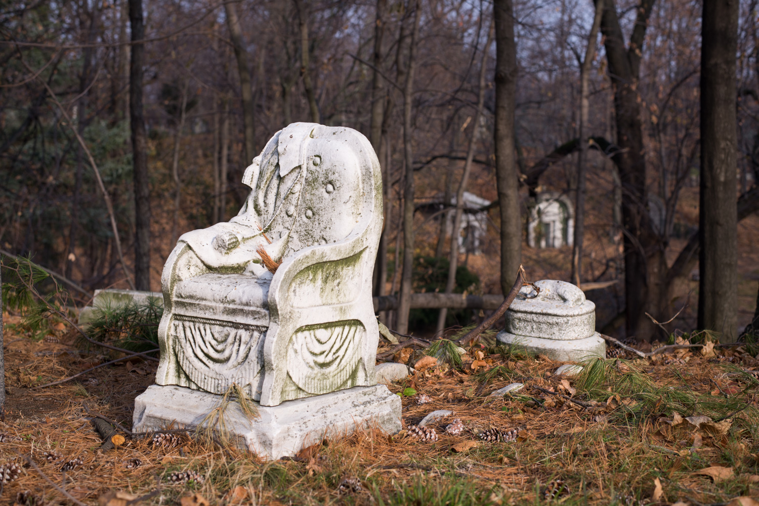 Color image of a weathered white marble throne statuary amongst brown needles and pine cones.