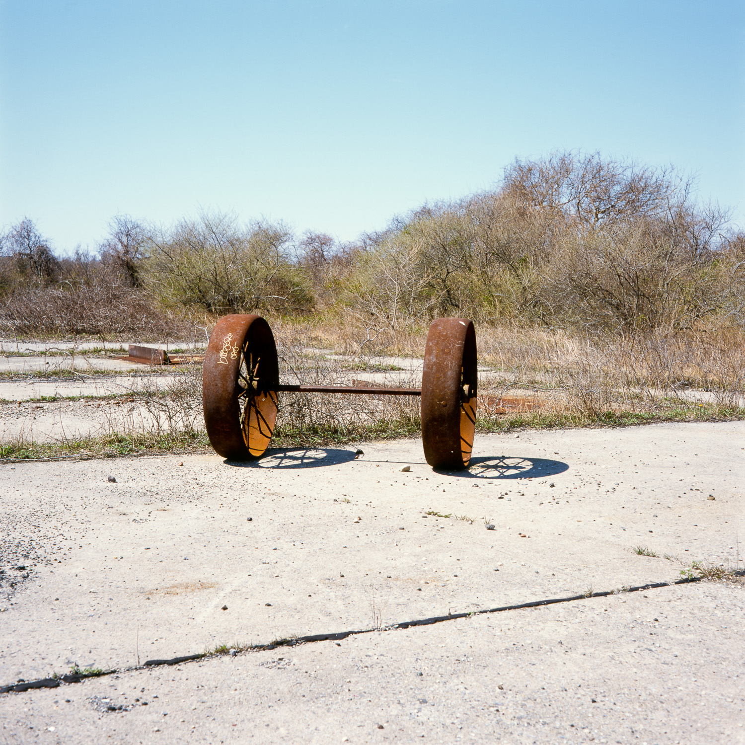 Color image of rusted set of wheels on an axle on a concrete foundation with weeds growing out of the cracks.