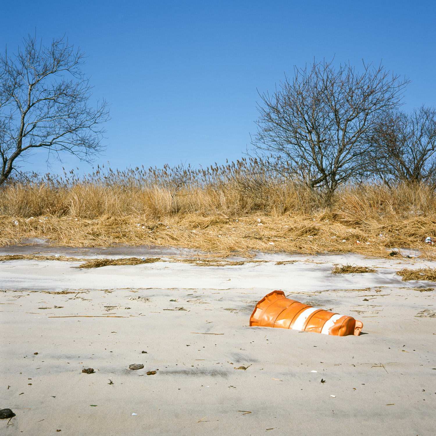 Color image of orange pylon half-buried in the sand of a beach with yellow grass behind it under a blue sky.