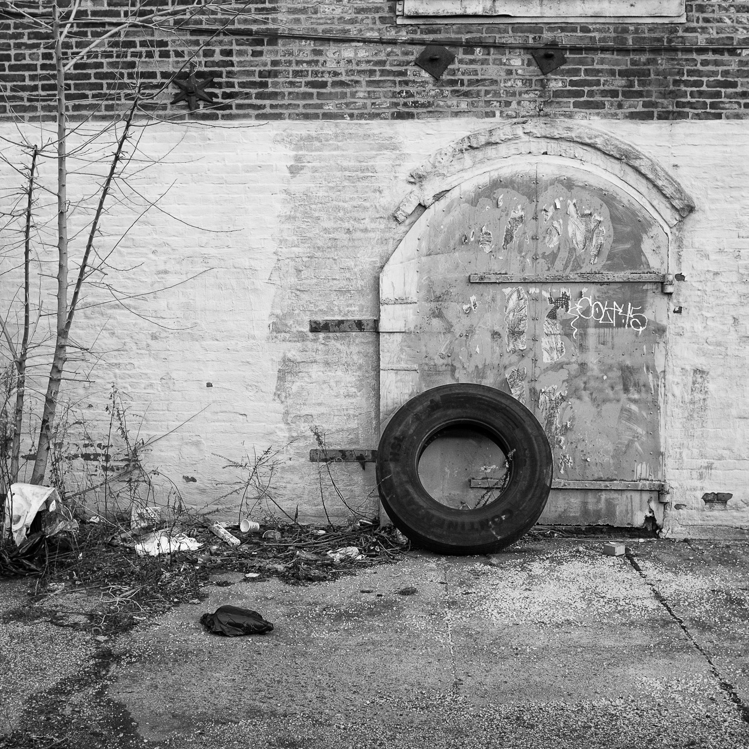 B&W image of tire against an old old iron door set into a white painted brick wall with remnants of torn off wheatpast art on wall