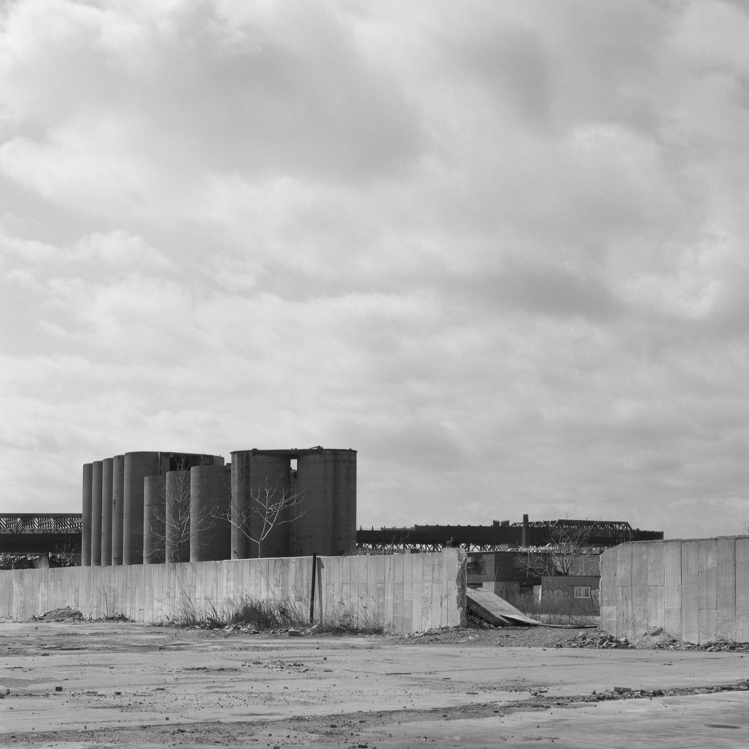 B&W image of an abandoned grain terminal in an empty concrete lot with an overhead train line in the distance.