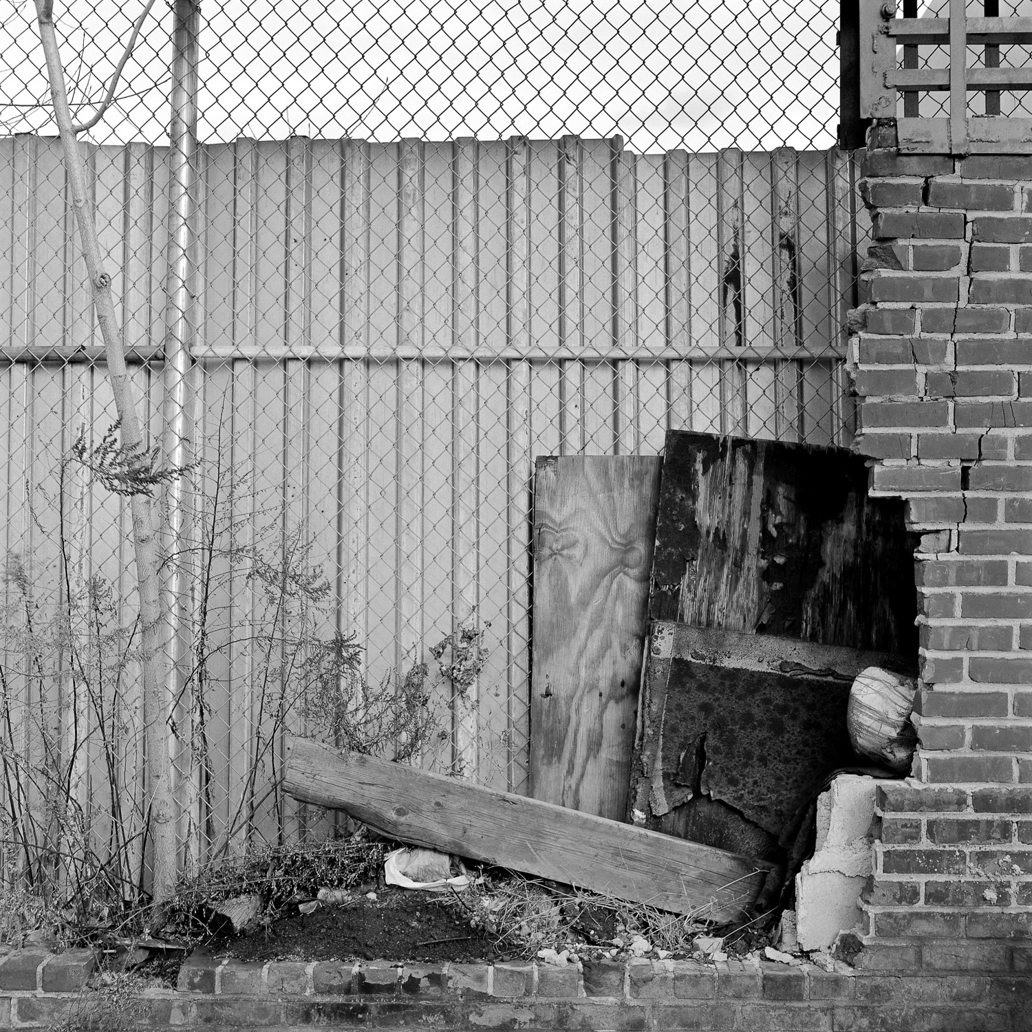 B&W image of chain link fence with brick wall and various pieces of debris wedged between them.