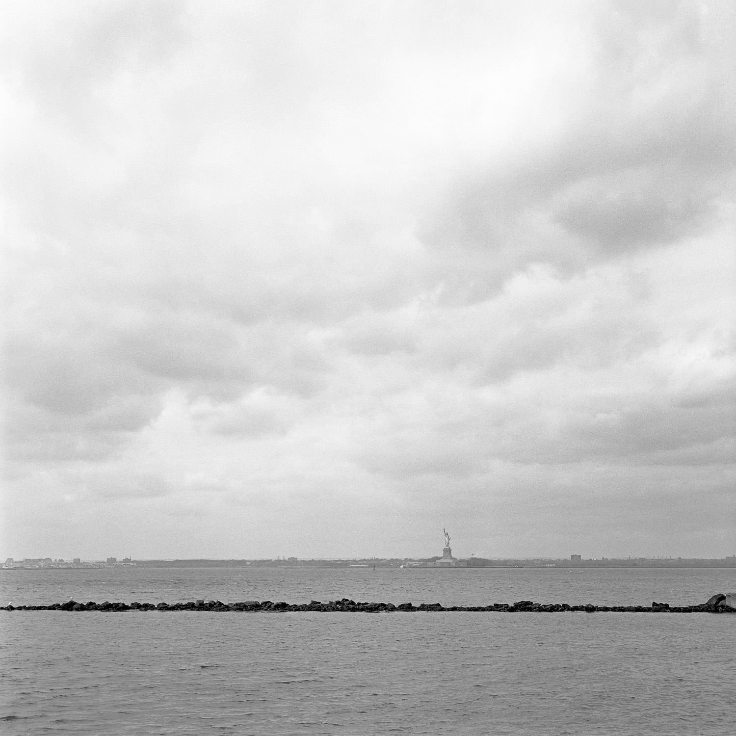 B&W image of a jetty in a sound under a cloudy sky with the Statue of Liberty in the distance.