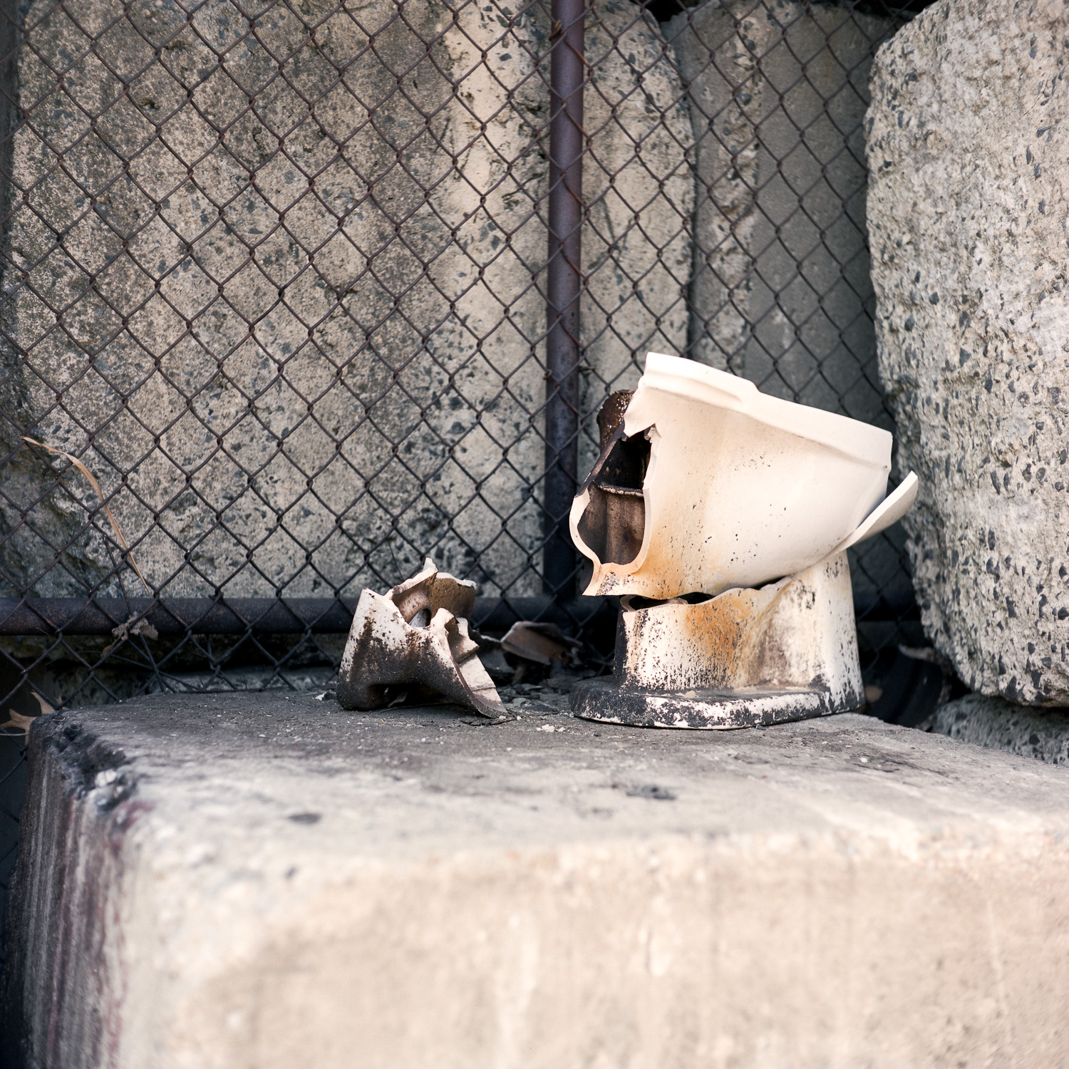 Color image of broken toilet on concrete block in front of black chain link fence.