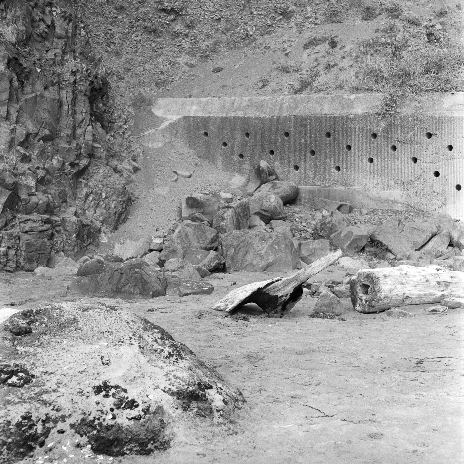 B&W image of a cement wall with holes in it on a cliff face wall on a beach.