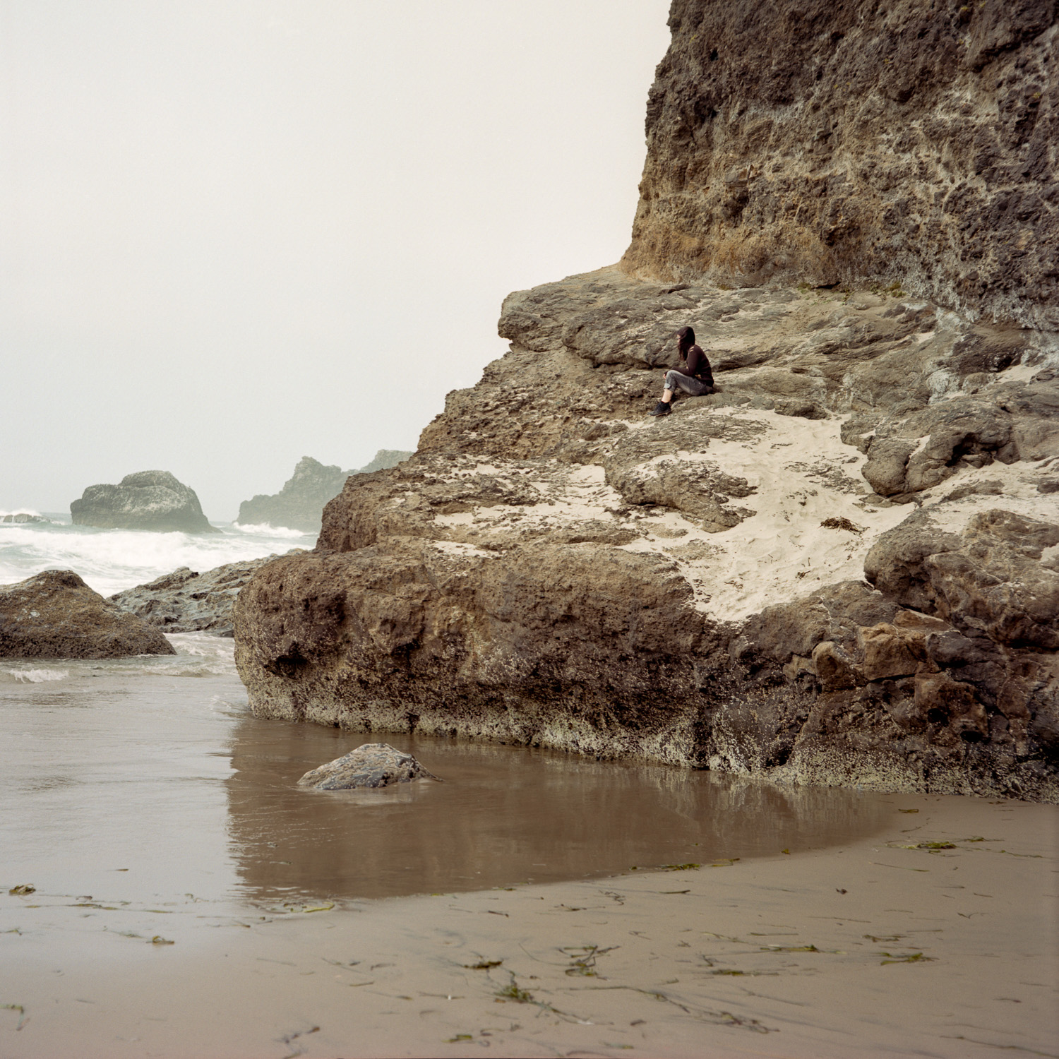 Color image of a lone figure sitting on a rock outcropping on the Pacific Ocean.