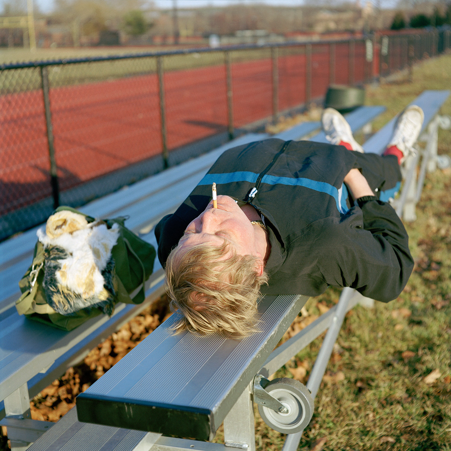 Color image of Siva avatar in a tracksuit smoking a cigarette laying down on a bleacher.